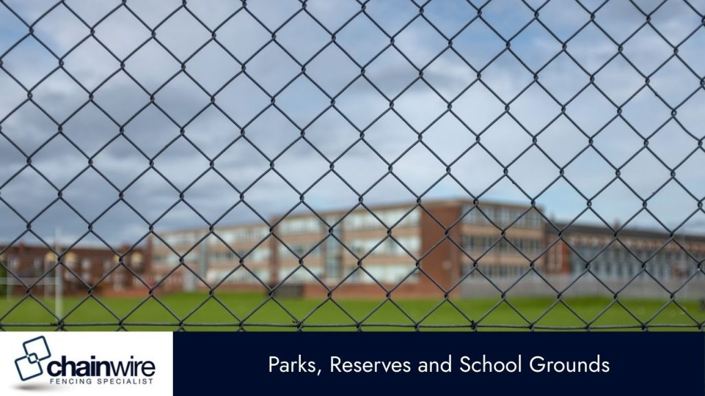 A close-up of a chain-link fence in the foreground, with a large, green sports field and a blurred school building behind it.