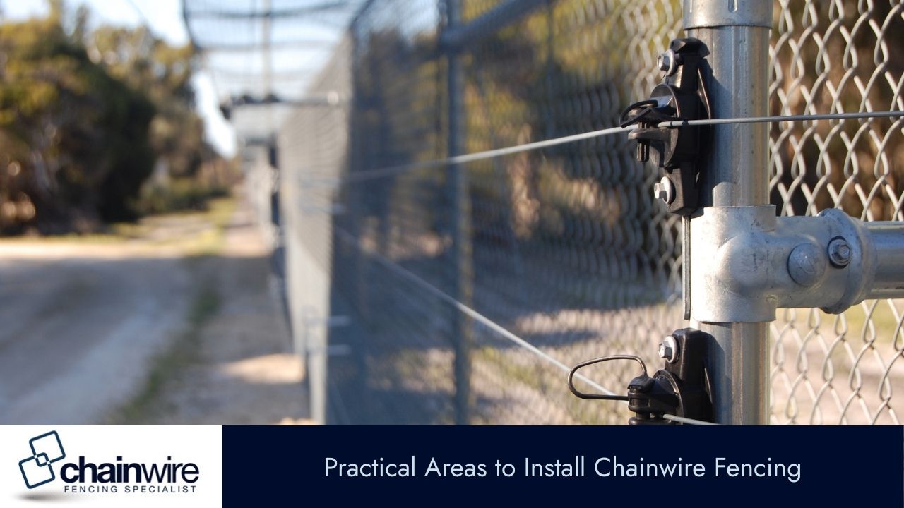 A close-up of a galvanized steel chainwire fence and post, with a dirt road and blurry foliage in the background.