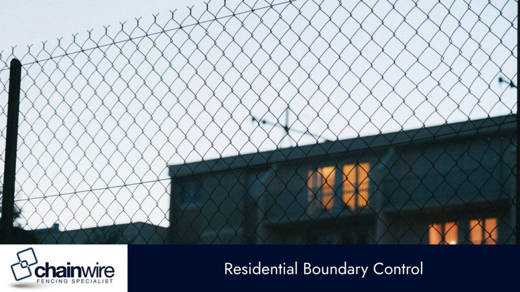 A close-up, silhouetted shot of a chain-link fence, with a house and lit windows blurred in the background. 