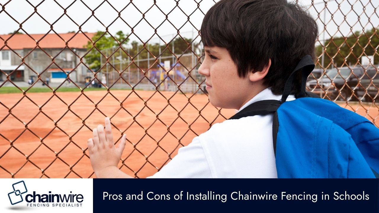 A boy with a backpack looks away while leaning his hand on a chain-link fence, which separates a school building and sports field.