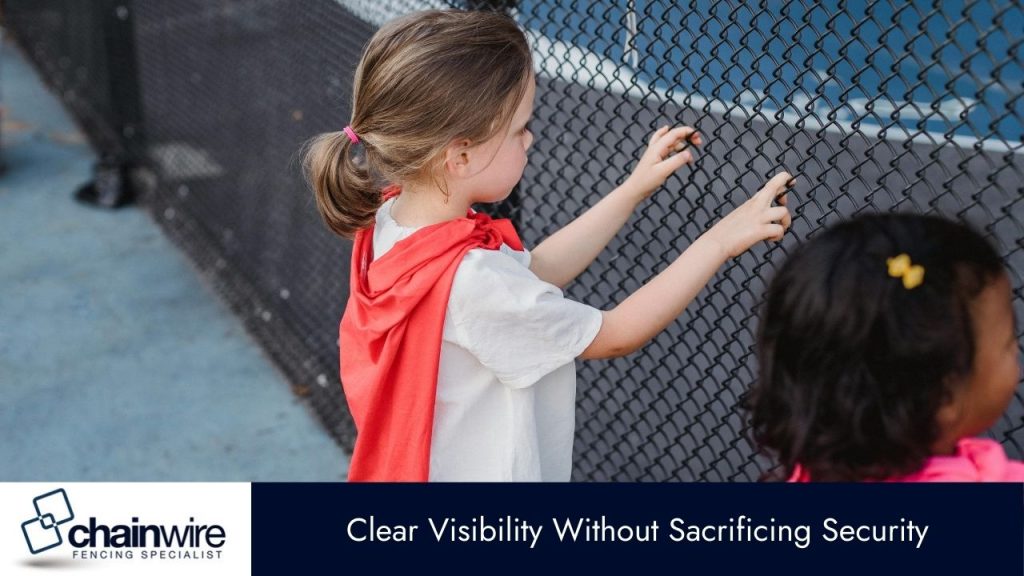 A young girl with a ponytail and a red cape is seen from the side, with her hands on a black chain-link fence. 