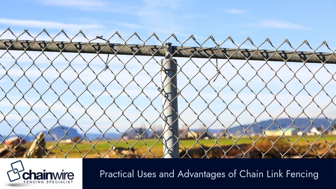 A close-up of a chain-link fence with a top rail, set against a backdrop of green fields and distant mountains.