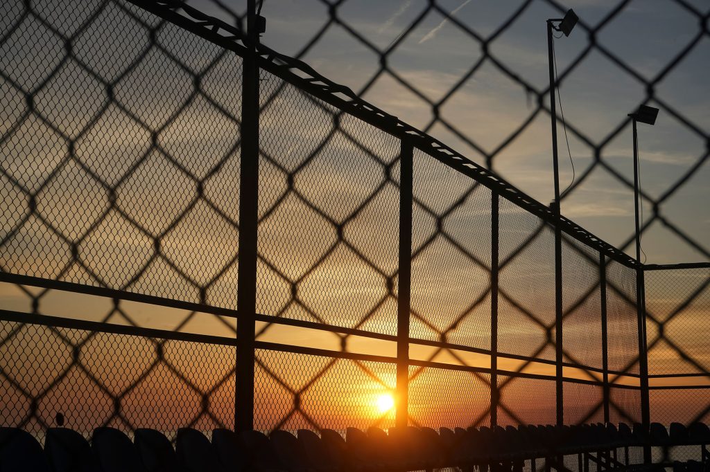 A chain-link fence is silhouetted against a vibrant sunset sky with hues of orange, yellow, and blue.