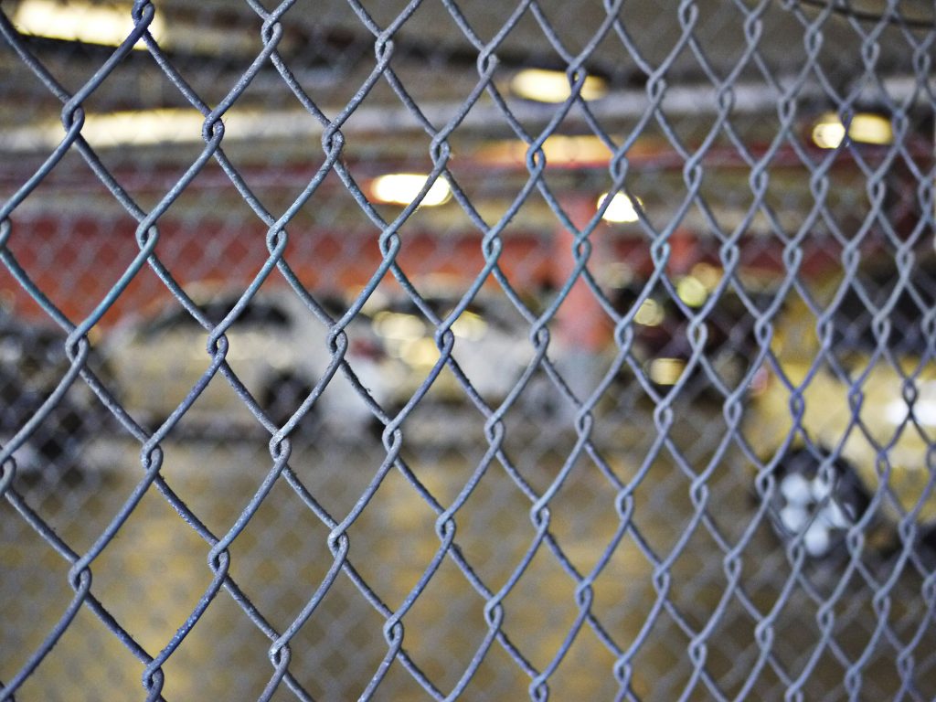 The background is blurred, showing a grey industrial or warehouse space with a car and other equipment behind the fence.