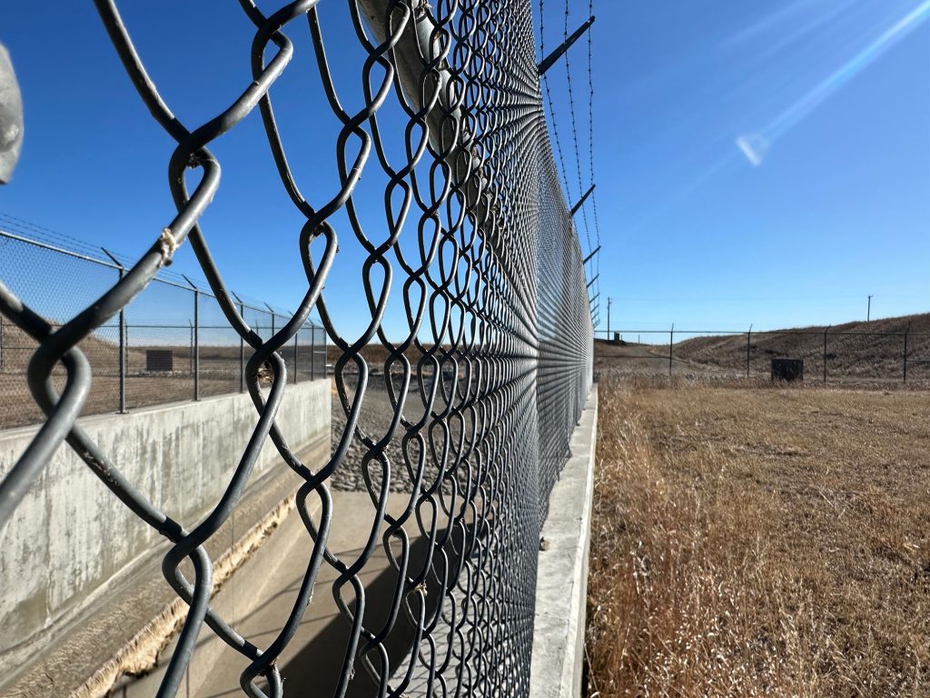 A side-angle view of a grey chain-link fence with a barbed wire top, set on a concrete base.