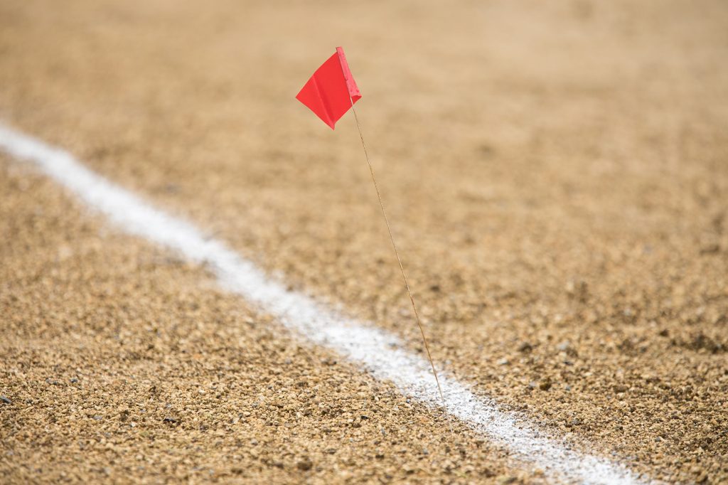 A close-up, low-angle shot of a small red flag on a thin wire stick, marking a spot in the dirt or sandy ground.