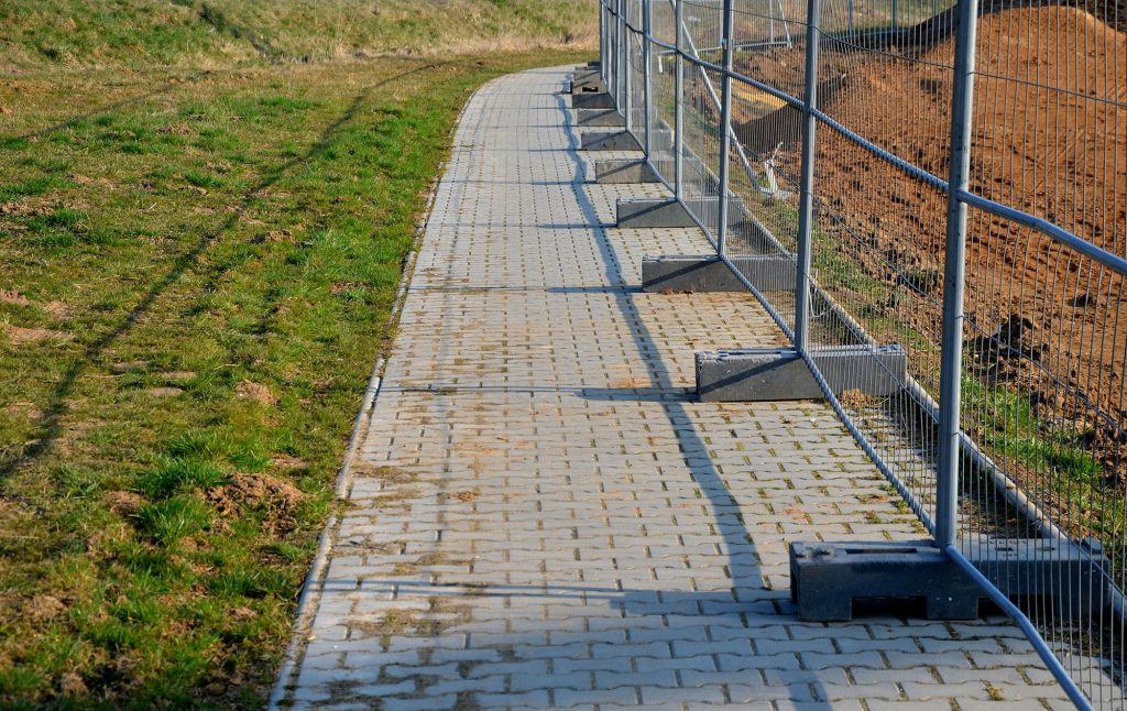 A paved walkway or path made of grey interlocking pavers, with a temporary metal barrier fence on the right side.