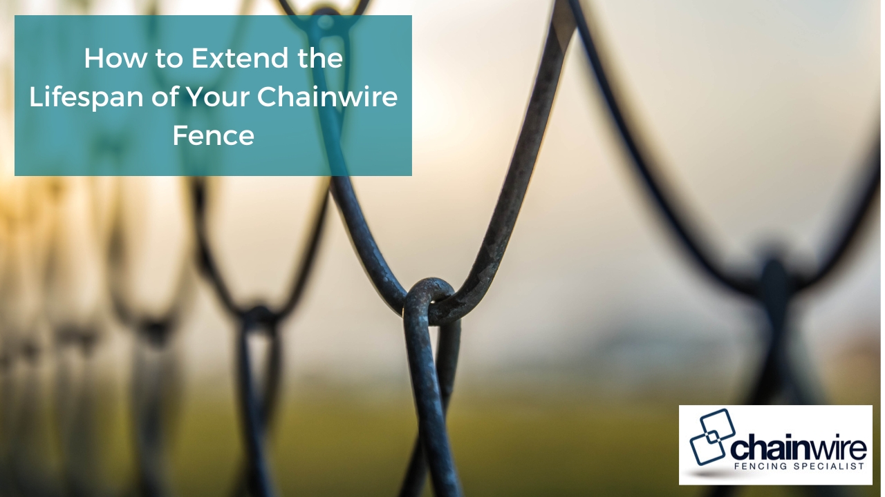 close-up of a chain link fence with a focus on the interweaving of the wires. The background is blurry, suggesting a field or outdoor setting. The wires are dark in color, possibly black or dark gray.