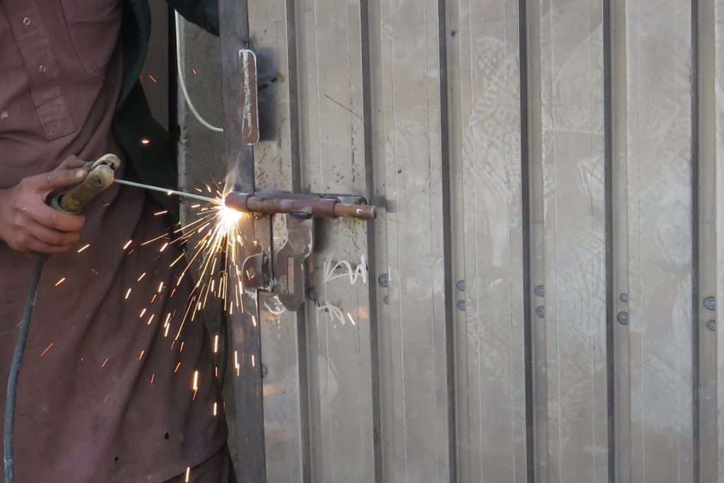 A welder, wearing brown work clothes, is using a welding torch to repair a large, heavy bolt lock on a metal gate.