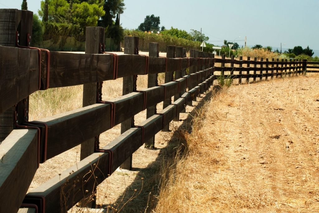 A long, rustic wooden fence, consisting of thick vertical posts and two horizontal rails, stretches into the distance along a dry, grassy dirt path.
